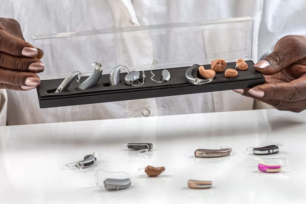 Hands with painted nails holding display of several hearing aids in front of lab coat with more hearing aids on white table below